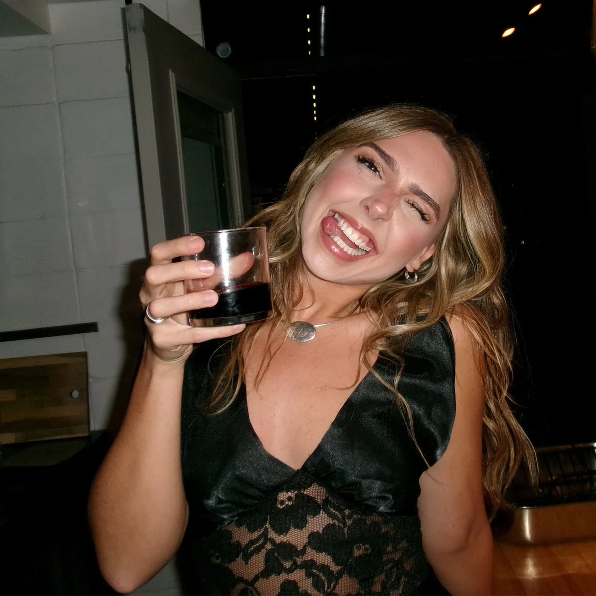 Woman holding a glass with a dark liquid, smiling in a casual setting.