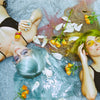 Two women lying on a gray concrete surface with water, white petals, and orange flowers around them. One has light blue hair and wears a black bikini top and dark sunglasses; the other has bright green hair and yellow-tinted sunglasses. taken with the Contax G1 Rangefinder Film Camera