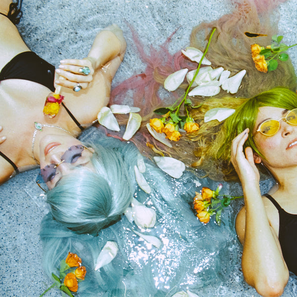 Two women lying on a gray concrete surface with water, white petals, and orange flowers around them. One has light blue hair and wears a black bikini top and dark sunglasses; the other has bright green hair and yellow-tinted sunglasses. taken with the Contax G1 Rangefinder Film Camera