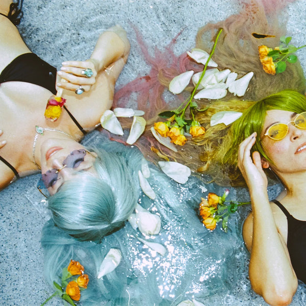 Two women lying on a gray concrete surface with water, white petals, and orange flowers around them. One has light blue hair and wears a black bikini top and dark sunglasses; the other has bright green hair and yellow-tinted sunglasses. taken with the Contax G1 Rangefinder Film Camera