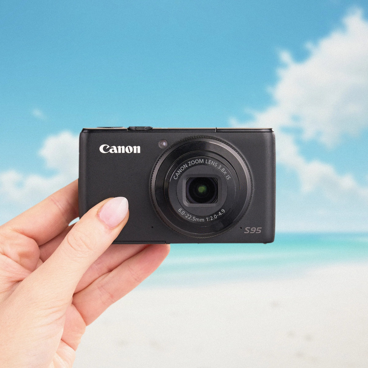 Canon PowerShot S95 compact camera held from the front against a beach backdrop