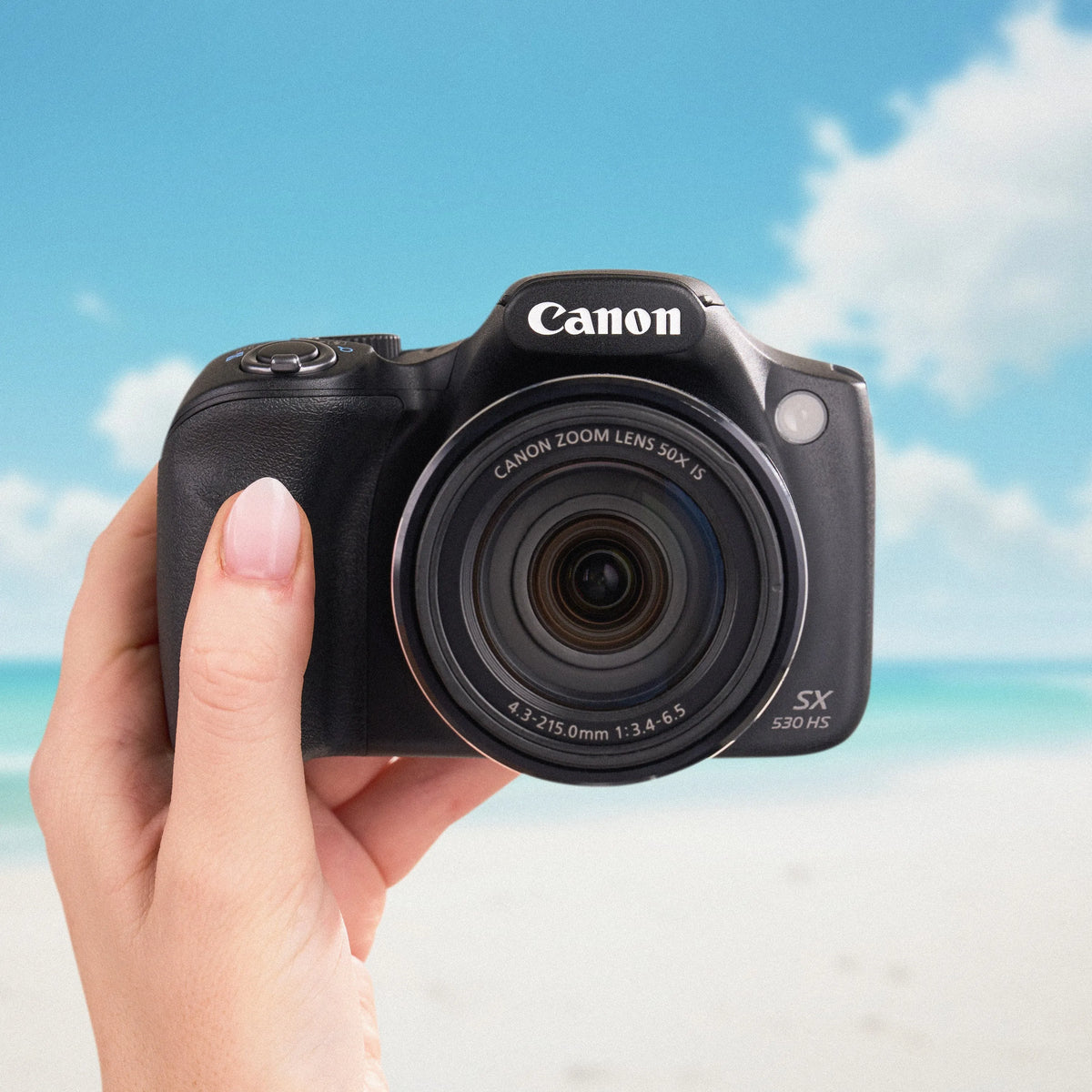 Canon PowerShot SX530 HS held from the front against a beach backdrop