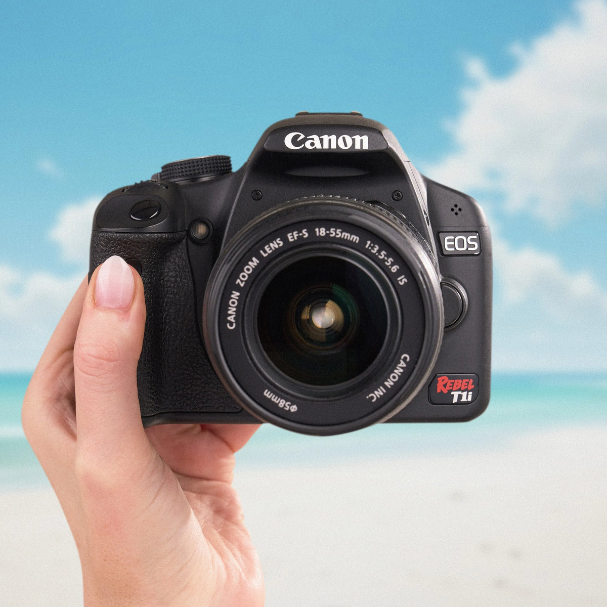 Canon Rebel T1i DSLR camera with an 18–55mm lens held against a beach backdrop
