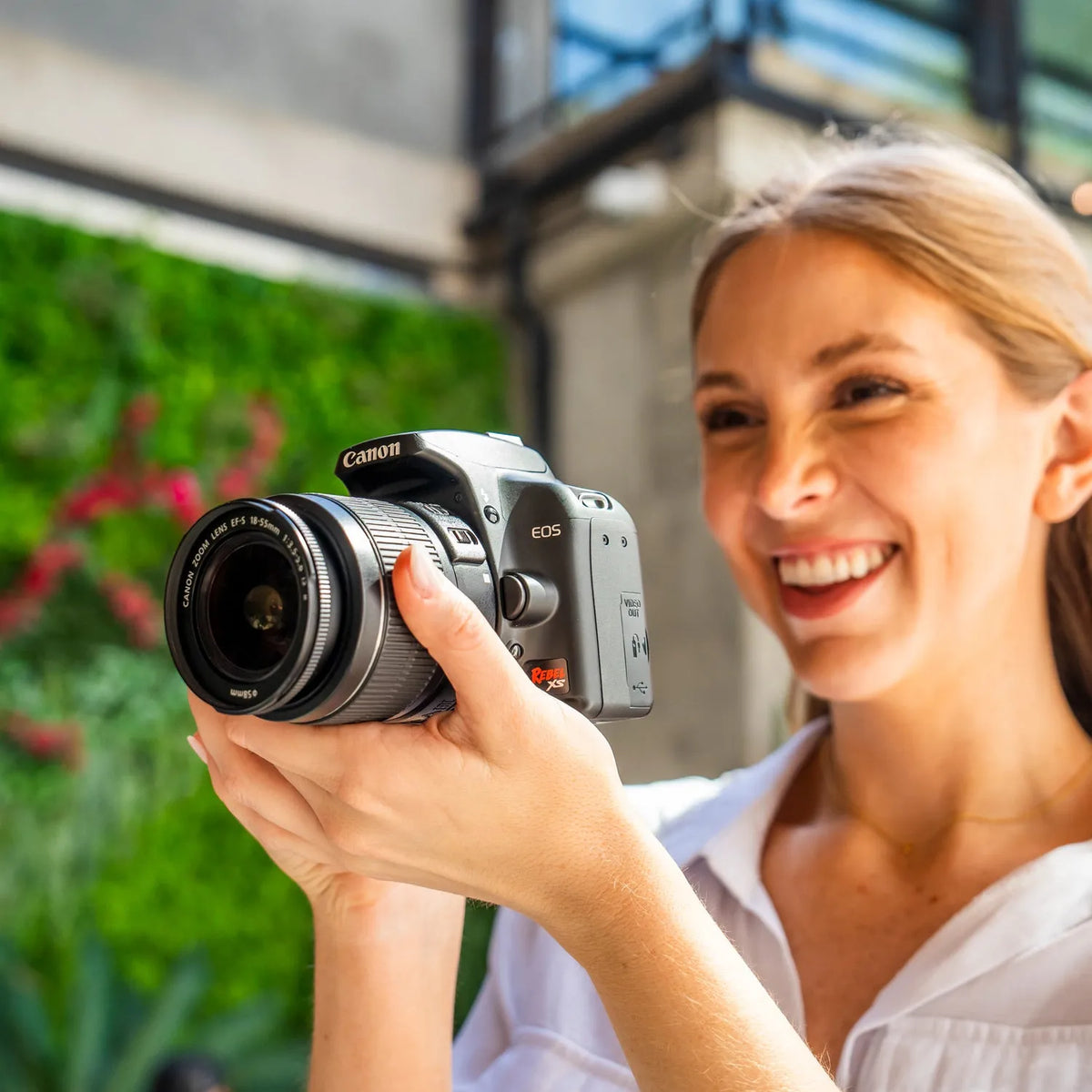 Woman holding a Canon Rebel XS with a blurred green and building background