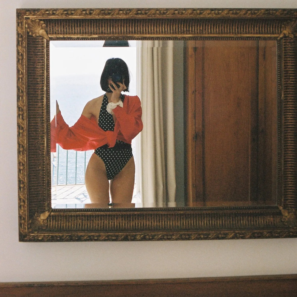 Person wearing a red shirt and black polka dot swimsuit reflected in an ornate mirror taken with the Nikon EM | 35mm Film Camera 