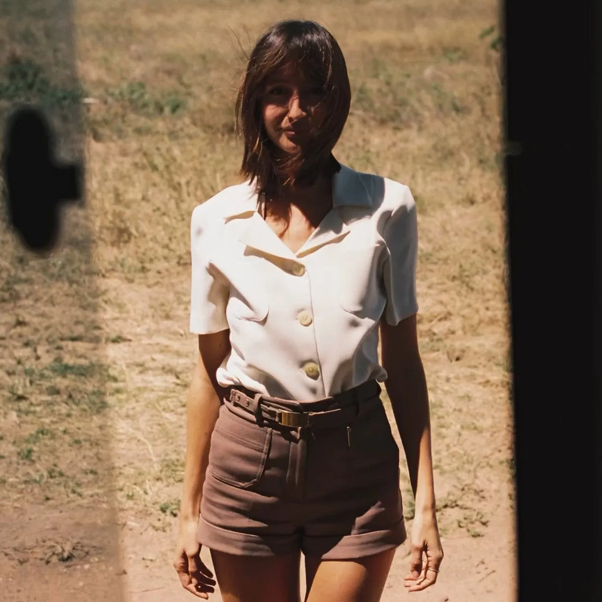 Woman wearing a white blouse with buttons and brown shorts standing in a dry, open field taken with a Nikon EM | 35mm Film Camera 