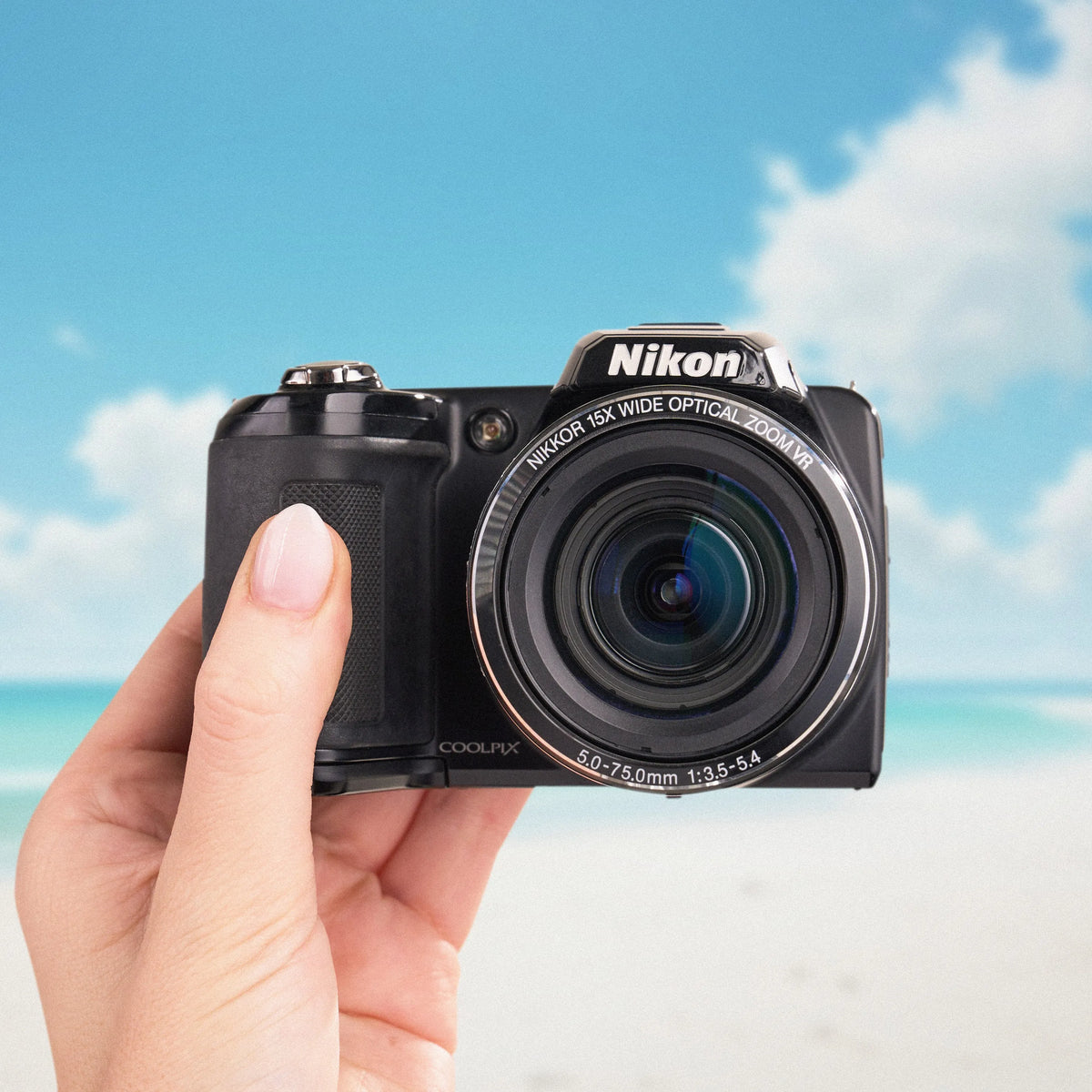 Front view of a black Nikon COOLPIX L110 held against a sunny beach background