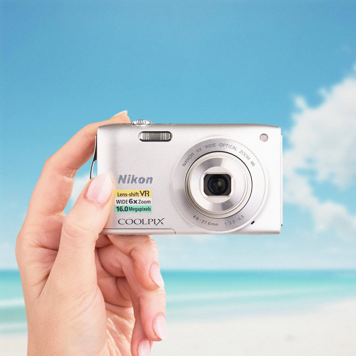 Front view of a silver Nikon COOLPIX S3300 held against a sunny beach background