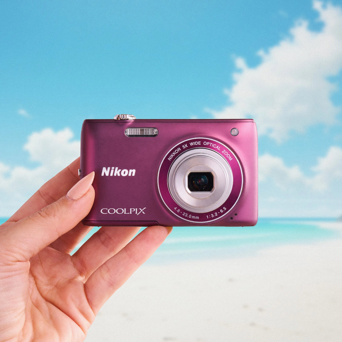 Front view of a purple Nikon Coolpix S4100 compact digital camera held against a beach background
