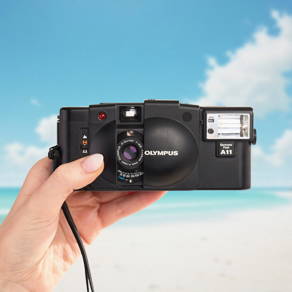 Hand holding an Olympus XA 2 | 35mm Point and Shoot Film Camera with a beach and blue sky in the background