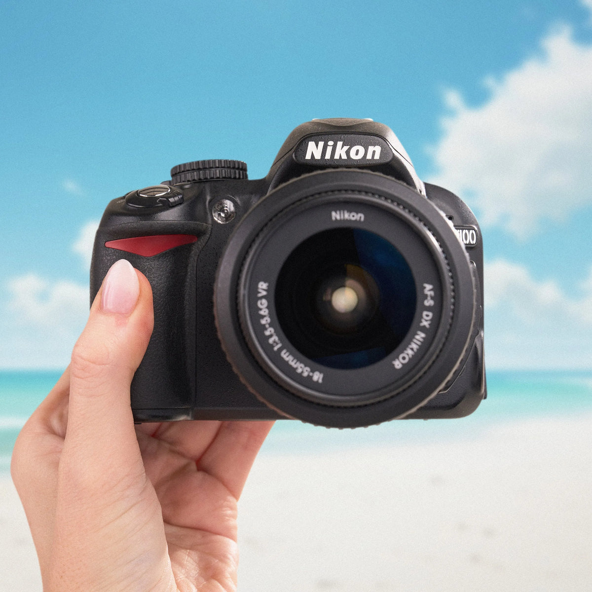 Front view of a black Nikon D3100 DSLR held against a sunny beach background