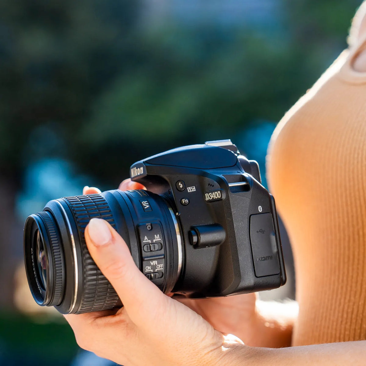 Close-up of a person holding a Nikon D3400 DSLR camera outdoors