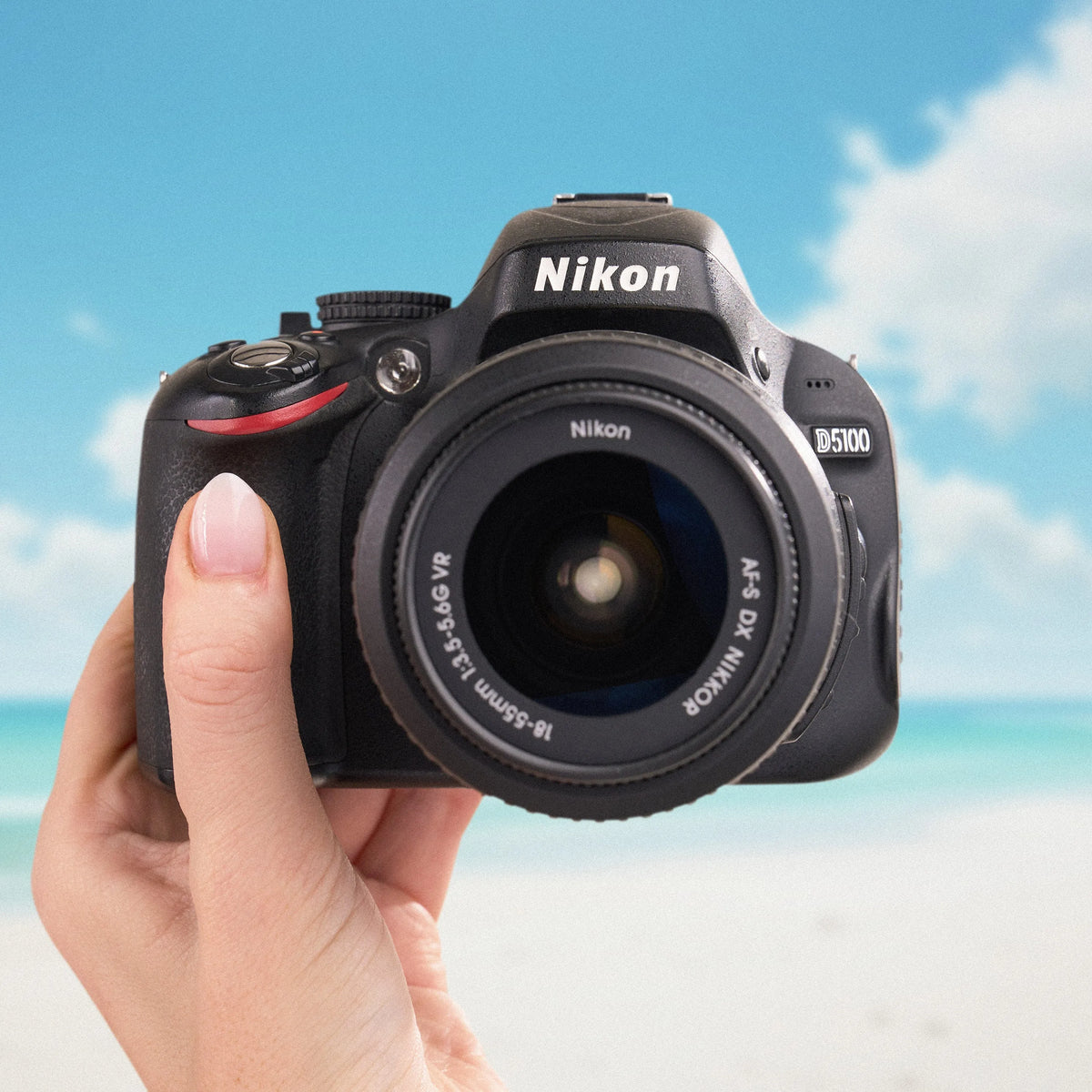 Nikon D5100 DSLR camera with an 18–55mm kit lens held in front of a bright beach scene