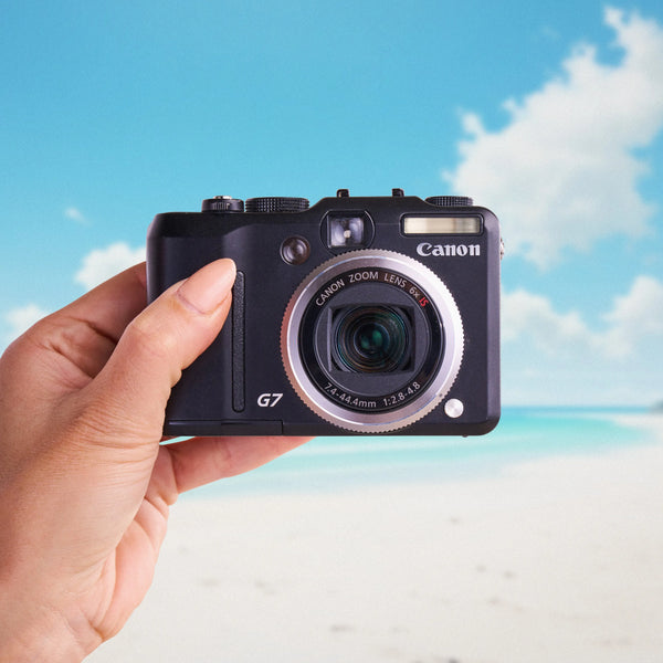 Front view of a Canon PowerShot G7 compact digital camera held against a beach background