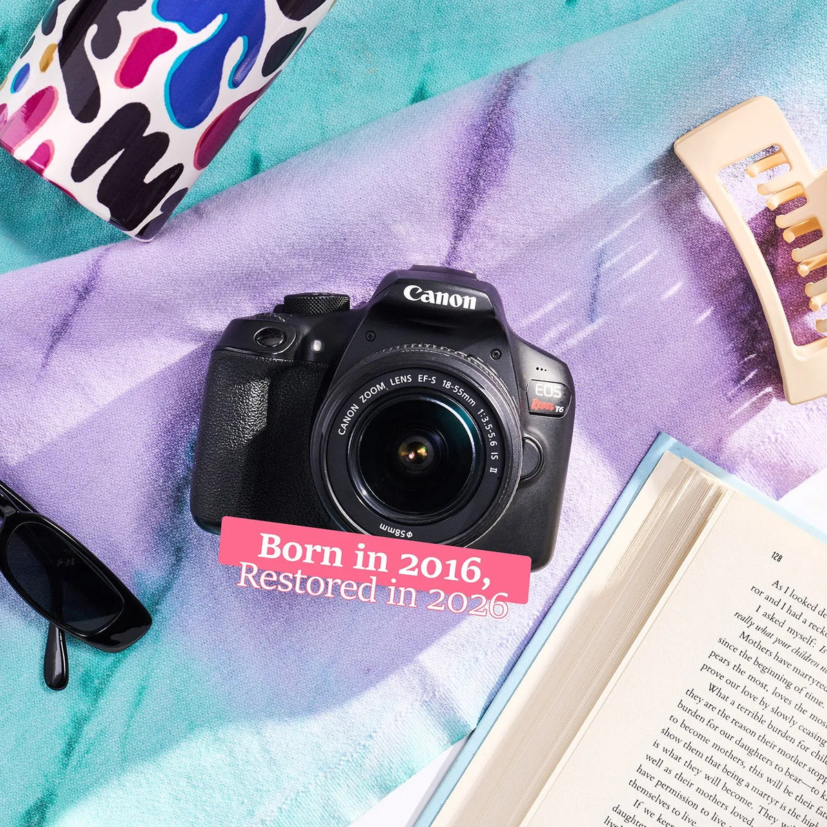 Canon Rebel T6 DSLR Camera placed on a purple and teal textured blanket with a pink text overlay reading 'Born in 2016, Restored in 2026'. Surrounding props include sunglasses, an open book, a colorful pouch, and a hair clip.