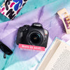 Canon Rebel T6 DSLR Camera placed on a purple and teal textured blanket with a pink text overlay reading 'Born in 2016, Restored in 2026'. Surrounding props include sunglasses, an open book, a colorful pouch, and a hair clip.