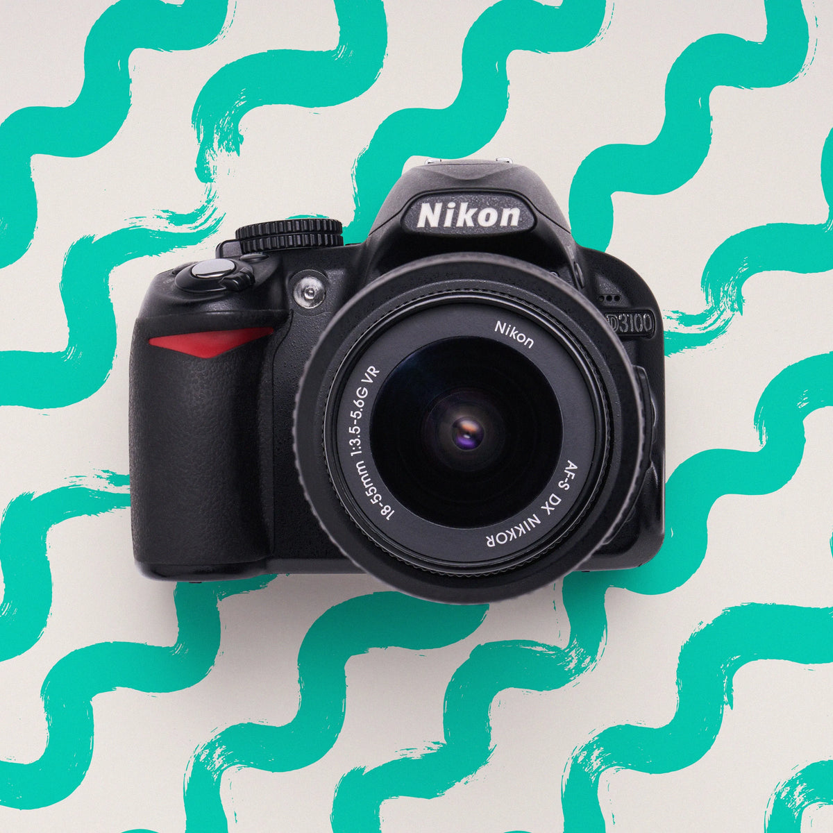Front view of a black Nikon D3100 DSLR camera with a NIKKOR lens on a teal wavy background