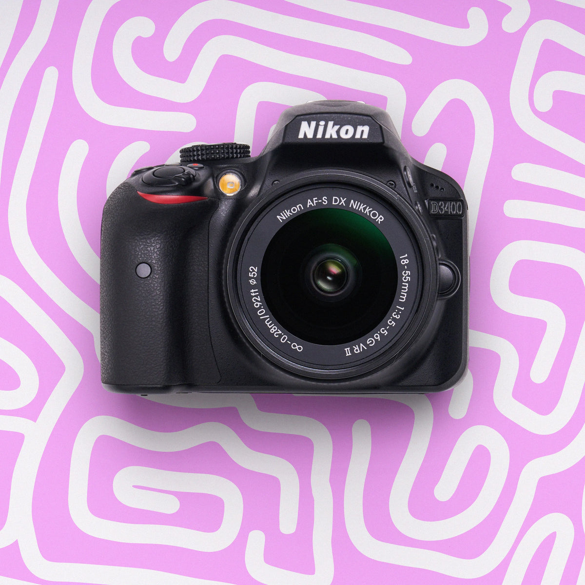 Front view of a Nikon D3400 beginner DSLR camera with an 18–55mm lens on a pink patterned background