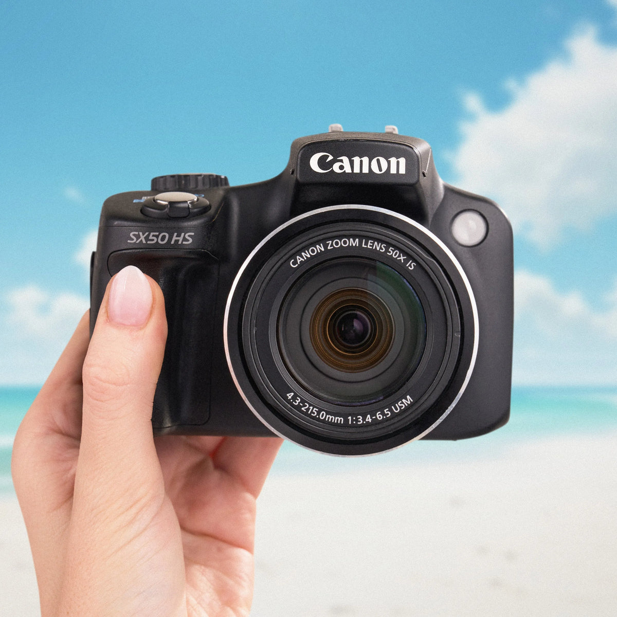 Front view of a black Canon PowerShot SX50 HS held against a sunny beach background