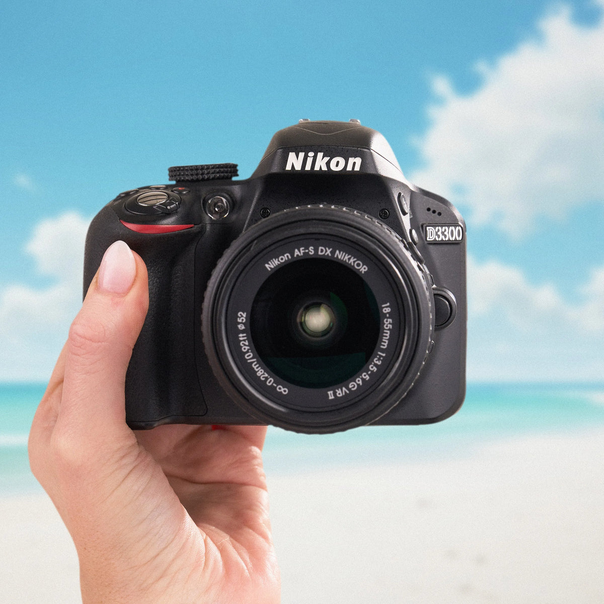 Front view of a Nikon D3300 DSLR with an 18–55mm kit lens held against a beach background
