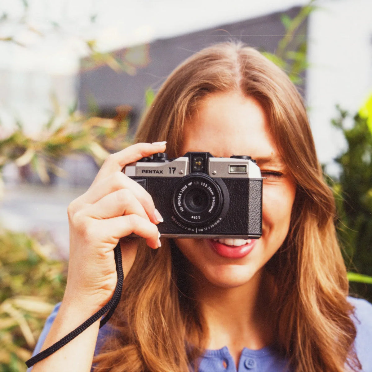 A woman with long light brown hair holds a Pentax 17mm camera in front of her face, smiling in an outdoor setting with blurred greenery and buildings in the background.