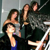 Four women posing together in glamorous outfits with a plain background