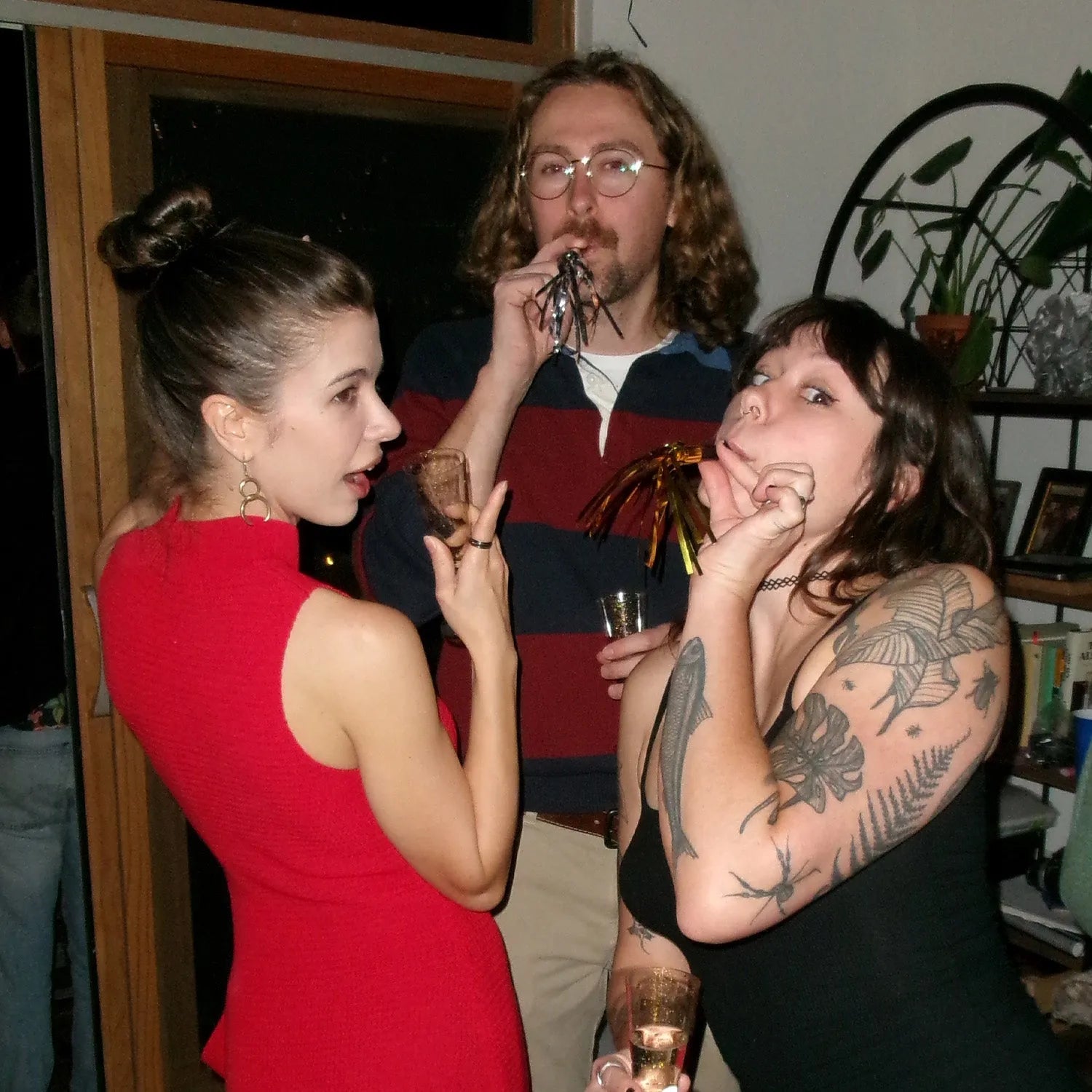 Two women and a man at a social gathering, with one woman in a red dress and the other with tattoos.