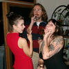 Two women and a man at a social gathering, with one woman in a red dress and the other with tattoos.