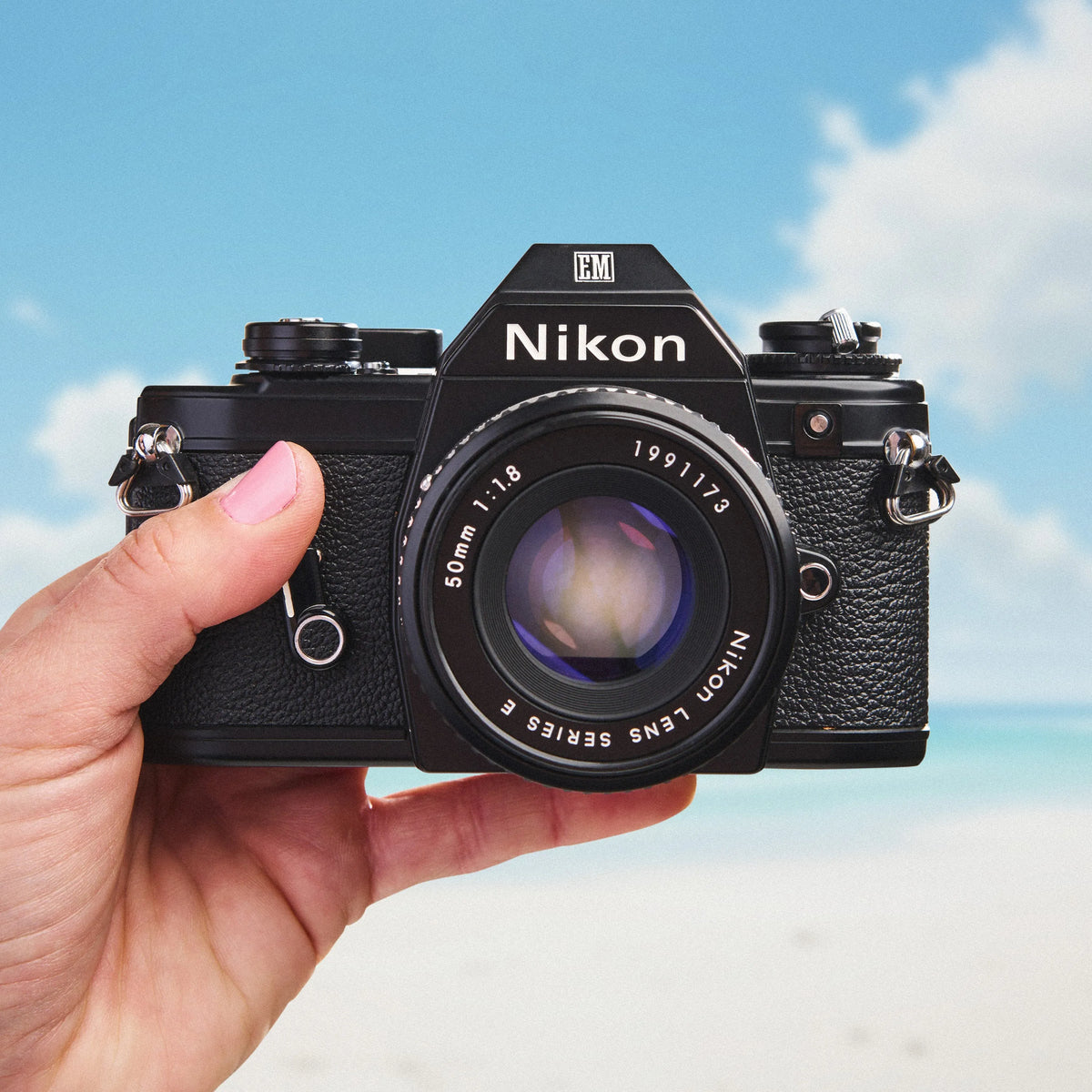 Hand holding a Nikon EM | 35mm Film Camera  against a blue sky background