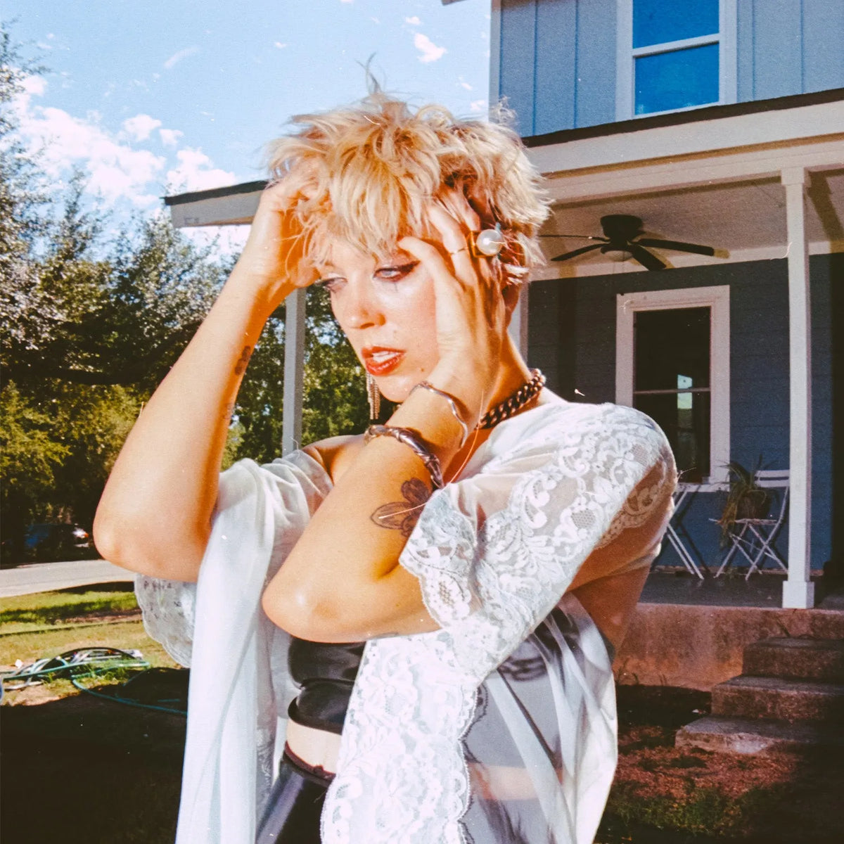 A woman with short blonde hair poses outdoors in front of a blue house, wearing a white lace long-sleeve top and a sheer white open-front cardigan. She has on multiple beaded necklaces and bracelets taken with the Nikon FM2 Film Camera