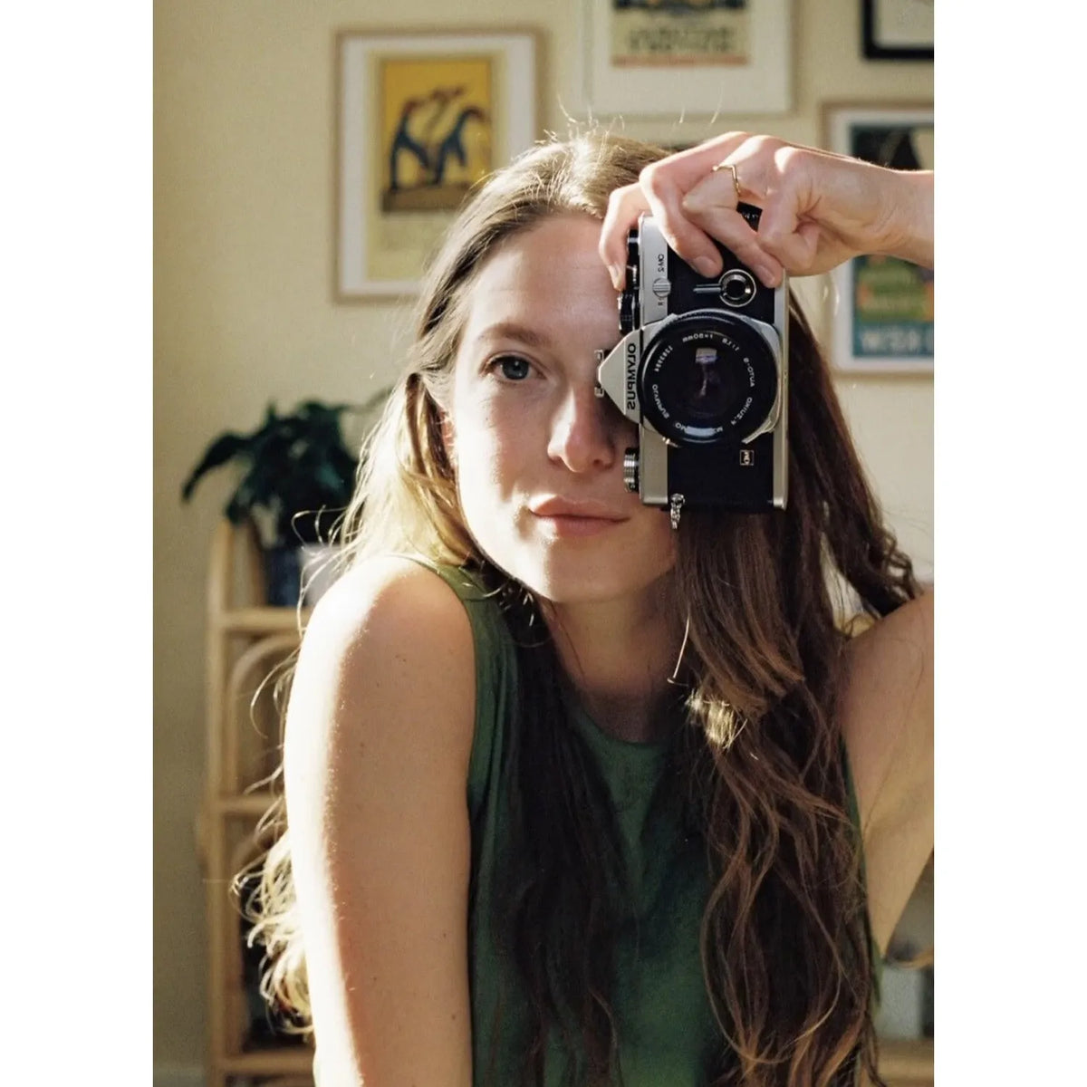 Woman holding a Olympus OM-1 | 35mm Film Camera up to her eye in a room with framed pictures on the wall.