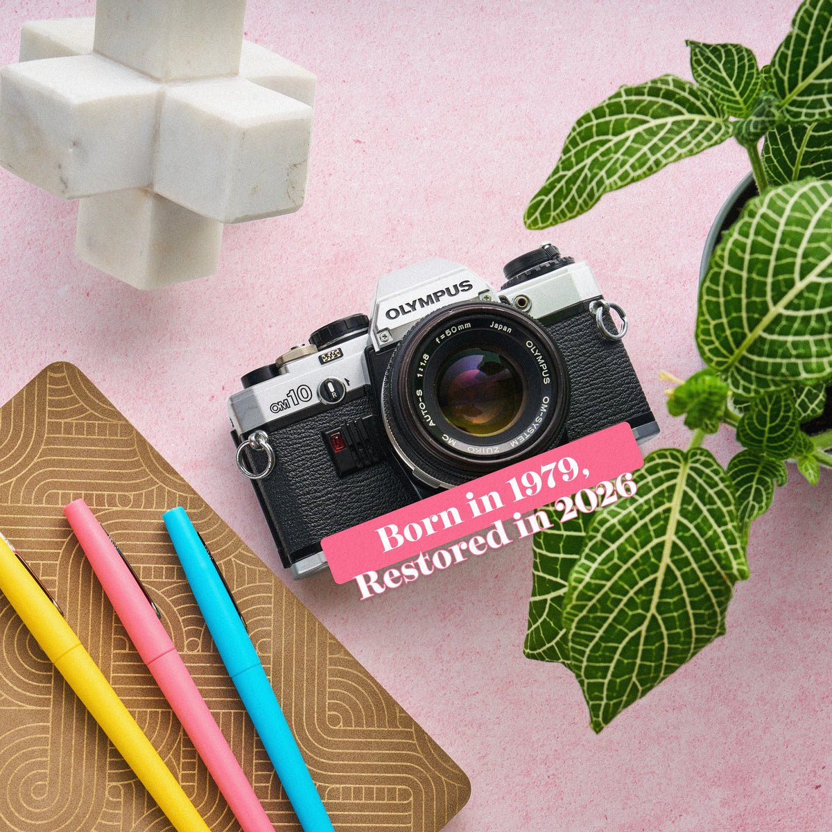 Olympus OM-10 35mm Film Camera  camera on a pink surface with stationery items and text overlay