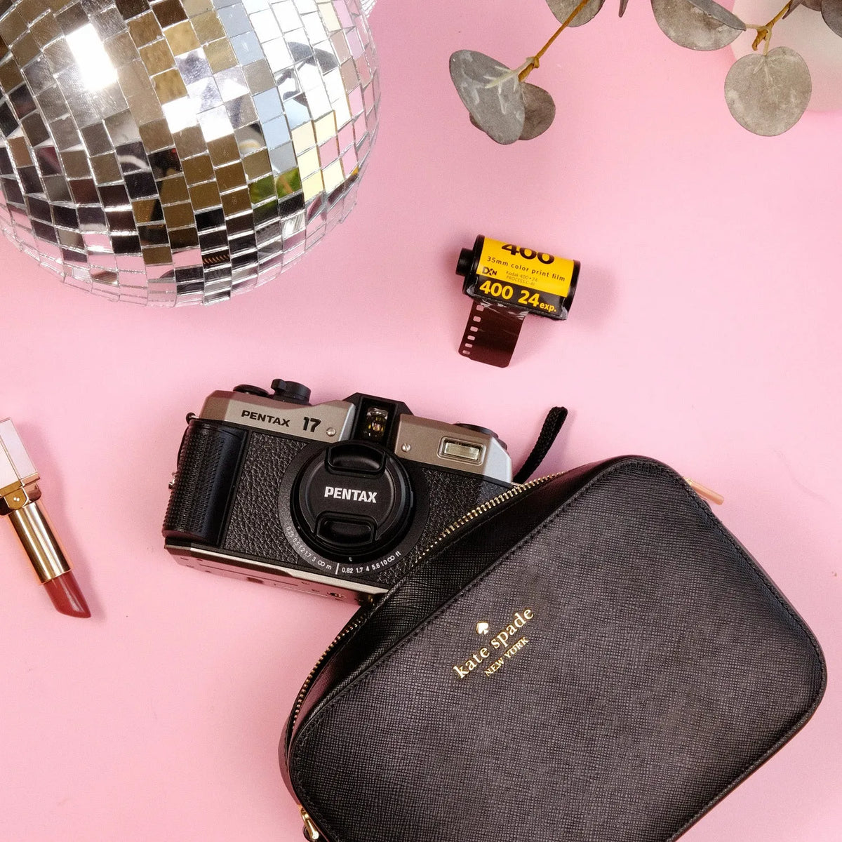 Pentax 17 Film Camera, film, and Kate Spade pouch on a pink background with disco ball and lipstick.
