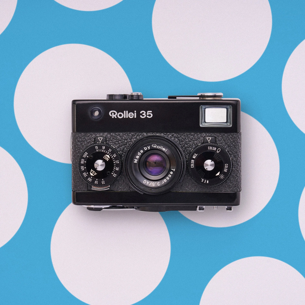 A vintage black Rollei 35 film camera photographed from above against a bright blue background with large white polka dots.