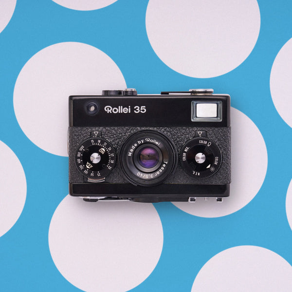 A vintage black Rollei 35 film camera photographed from above against a bright blue background with large white polka dots.